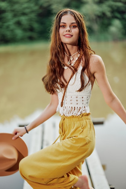 Een jonge vrouw in een hippie-look en ecodress danst buiten bij het meer met een hoed en een gele broek op in de zomerzonsondergang