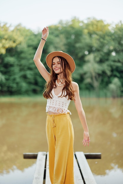 Een jonge vrouw in een hippie-look en ecodress reist door de natuur bij het meer met een hoed en een gele broek op in de herfstzonsondergang