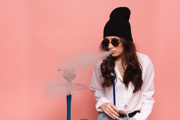 Een jonge vrouw in een hipster-afbeelding rookt een waterpijp, sheesha en geniet van roken.