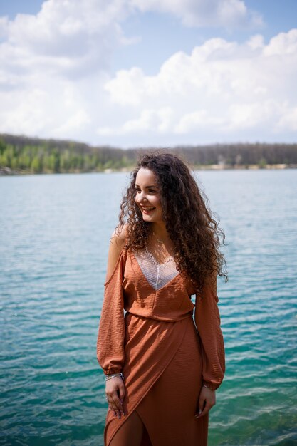 Een jonge vrouw in een jurk staat op houten metselwerk in het midden van een blauw meer. Meisje gelukkig glimlacht en de zon schijnt, zomerdag. Ze heeft lang krullend haar en een Europese uitstraling