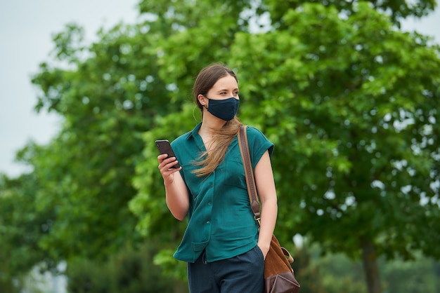 Een jonge vrouw in een marineblauw medisch gezichtsmasker leest nieuws op een smartphone en steekt een hand in een zak broek lopen