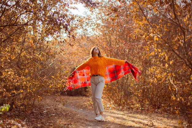 Een jonge vrouw in een oranje trui wikkelt zichzelf in een sjaal in een herfstpark