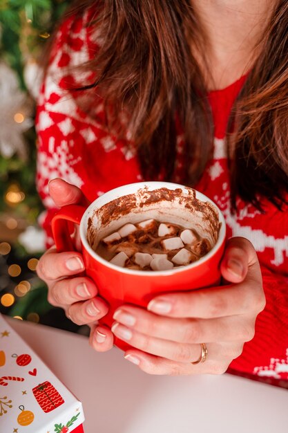 Een jonge vrouw in een rode trui houdt cacao met marshmallows vast naast een kerstboom