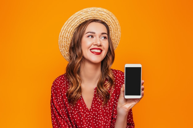 Een jonge vrouw in een rode zomerjurk met strohoed houdt een mobiele telefoon vast en laat deze zien aan de camera met een leeg zwart scherm en kijkt naar de mockup-ruimte voor een oranje ontwerp