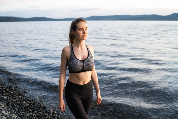 Een jonge vrouw in een sportbeha en legging loopt tijdens zonsondergang langs de rotsachtige oever van het Turgoyak-meer