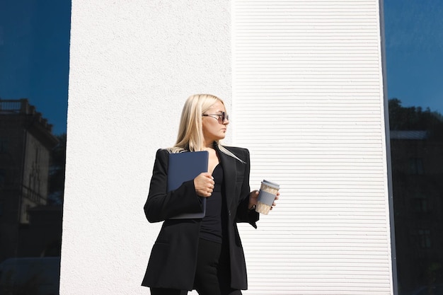 Een jonge vrouw in een stijlvol pak met een laptop en een kopje koffie poseert tegen de achtergrond van een zakencentrum