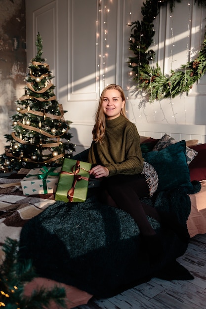 Een jonge vrouw in een trui opent kerstcadeaus in een slaapkamer die is ingericht voor kerstmis tegen de achtergrond van een kerstboom.