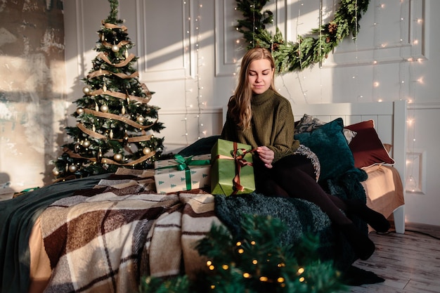 Een jonge vrouw in een trui opent kerstcadeaus in een slaapkamer die is ingericht voor Kerstmis tegen de achtergrond van een kerstboom.