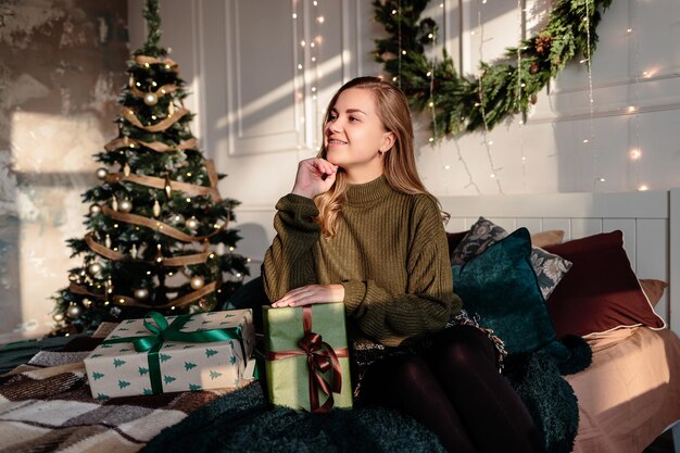 Een jonge vrouw in een trui opent kerstcadeaus in een slaapkamer die is ingericht voor Kerstmis tegen de achtergrond van een kerstboom.