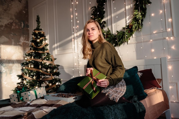 Een jonge vrouw in een trui opent kerstcadeaus in een slaapkamer die is ingericht voor kerstmis tegen de achtergrond van een kerstboom