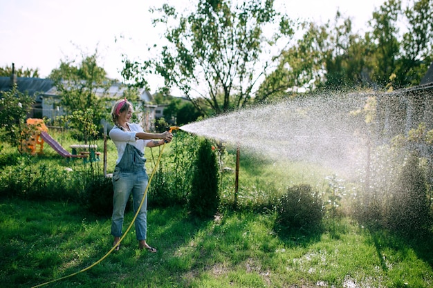 een jonge vrouw in een wit overhemd en een spijkerbroek die het gazon water geeft