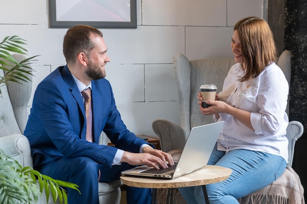 Een jonge vrouw in een wit overhemd en een spijkerbroek met een mok koffie en een Arabische man van middelbare leeftijd in een pak pratend aan een cafétafel met een laptop