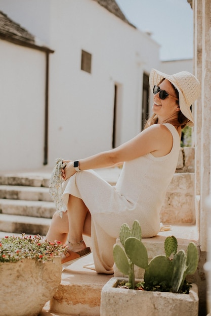 Een jonge vrouw in een witte jurk en hoed ontspant zich op de trap op een zonnige dag tijdens een toeristisch bezoek in Alberobello Italië