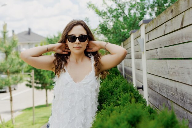 Een jonge vrouw in een witte jurk en zonnebril poseert tegen de achtergrond van een plattelandsdorpje Goed humeur en zomerse sfeer