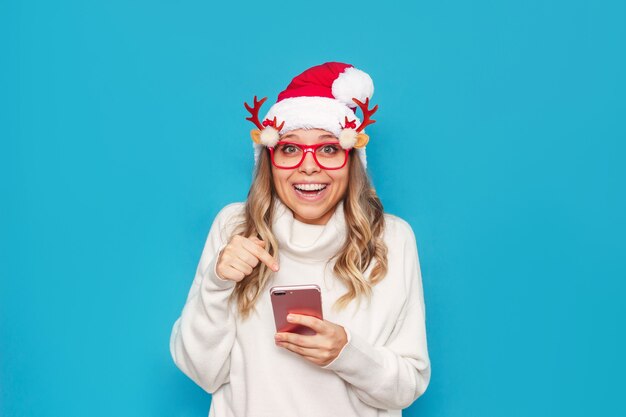 Een jonge vrouw in een witte trui kerstmuts kerstbril wijst naar het scherm van een mobiele telefoon