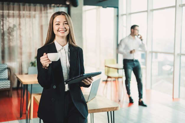 Een jonge vrouw in een zwart klassiek pak drinkt koffie terwijl ze in een modern kantoor staat. Werk in het kantoor op een draagbare tablet.