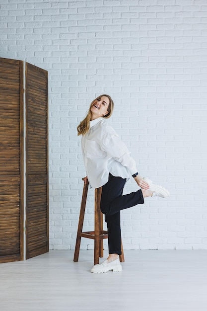 Een jonge vrouw in een zwarte broek en een wit overhemd tegen een witte bakstenen muur een stijlvol modern meisje in witte leren loafers