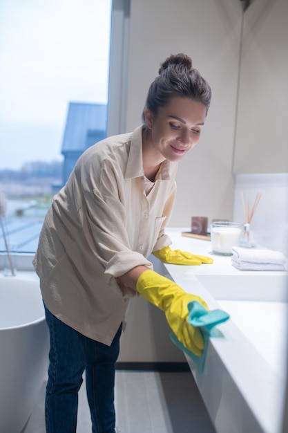 Een jonge vrouw in gele handschoenen die de oppervlakken in de badkamer schoonmaakt