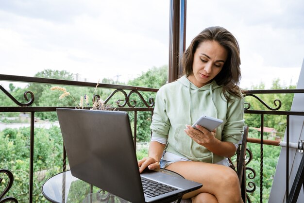 Een jonge vrouw in huiskleding zit op het balkon aan een tafel en werkt met een telefoon op een laptop in de s...