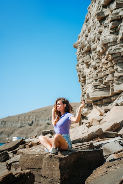 Een jonge vrouw in korte broek zit in een yogahouding op een rotsachtig strand gemaakt van natuursteen op de Krim