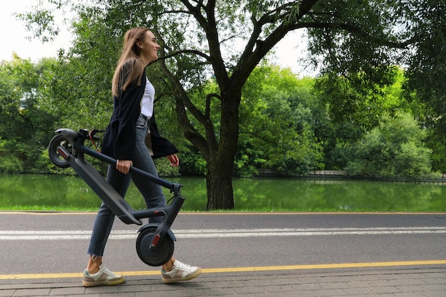 Een jonge vrouw in stijlvolle kleding loopt langs een fietspad met een opgevouwen elektrische scooter