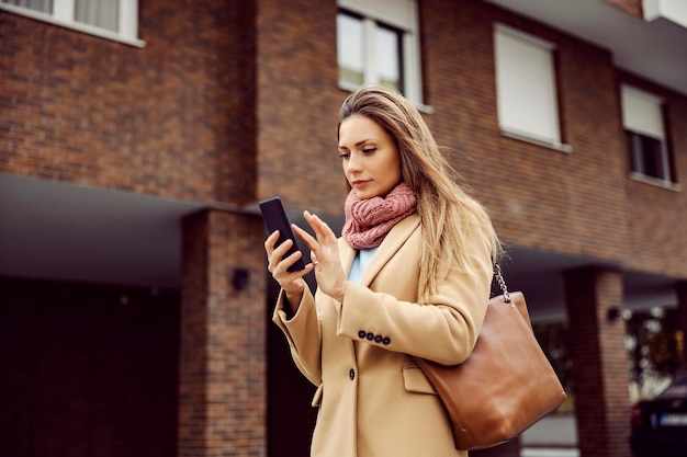 Een jonge vrouw in warme kleren die bij koud, winderig weer op straat staat en aan het typen is aan de telefoon