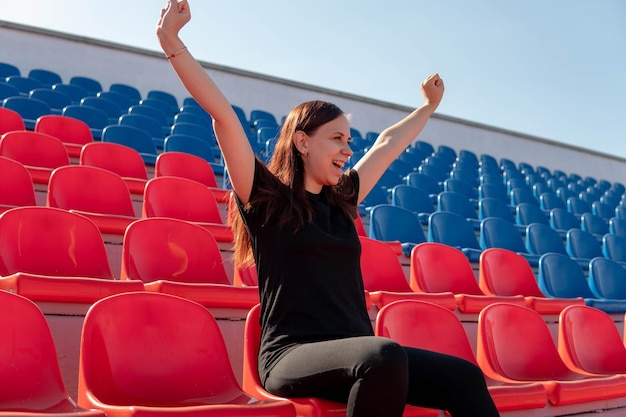 Een jonge vrouw in zwarte kleding met lang haar zit alleen op de tribunes van een stadion en steunt een favoriet sportteam voor haar