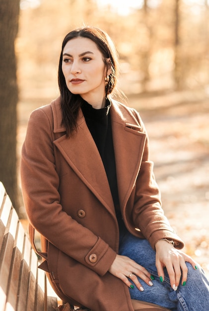 Een jonge vrouw is een bruette die in de herfst in een jas op een bankje in het park zit