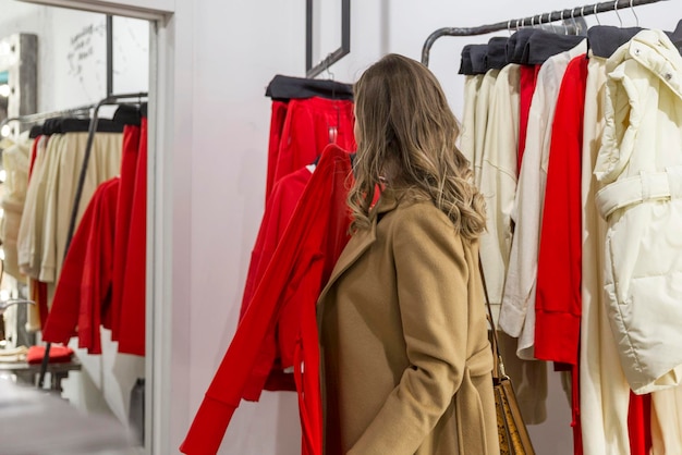 Een jonge vrouw kiest kleding in een modewinkel Een mooie blonde in een stijlvolle beige jas kijkt naar jurken op hangers Winkelen en vrije tijd