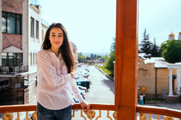 Een jonge vrouw kijkt uit vanaf het balkon. De felle zon verlicht het meisje. Mooie brunette geniet van de zon.