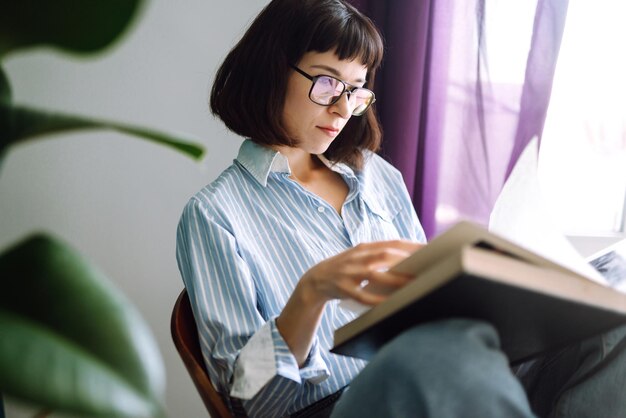 Een jonge vrouw leest een boek thuis, zittend in haar woonkamer. Concept van rust, ontspanning.