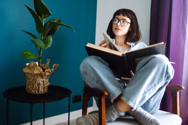 Een jonge vrouw leest een boek thuis, zittend in haar woonkamer. Concept van rust, ontspanning.