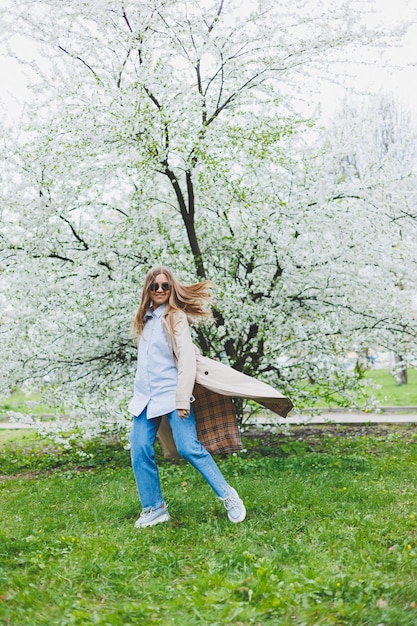 Een jonge vrouw met blond haar geniet van een bloeiende lentetuin Reizen voorjaarsvakantie Modieuze stijl Een vrouw in zonnebril en een beige trenchcoat loopt door een bloeiend park Selectie focus