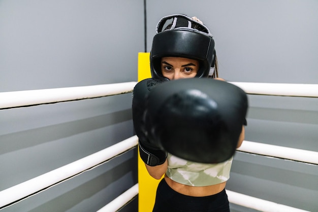 Een jonge vrouw met bokshandschoenen en beschermende helm gooit een klap naar de camera op een boksring