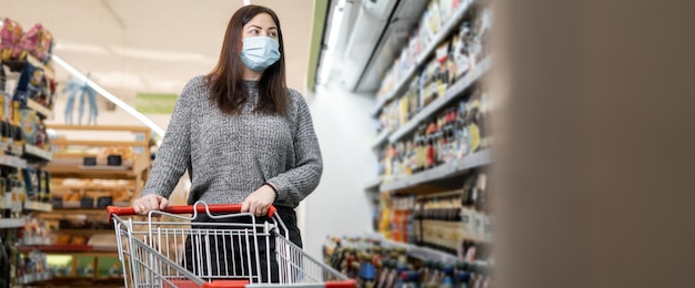 Een jonge vrouw met een beschermend masker in een supermarkt loopt met een winkelwagentje en selecteert producten in de schappen Supermarkt en shoppers Plaats voor uw tekst