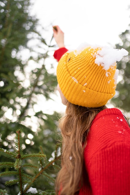 Foto een jonge vrouw met een besneeuwde hoed reikt naar een kerstboomtak en heeft plezier buitenshuis