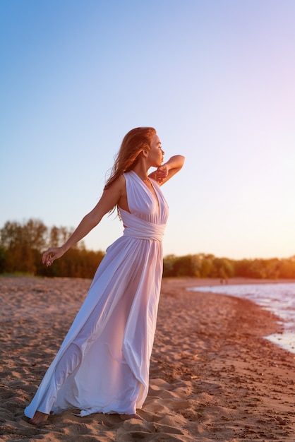 Een jonge vrouw met een europees uiterlijk in de zomer in een witte jurk aan de kust staat bij zonsondergang...