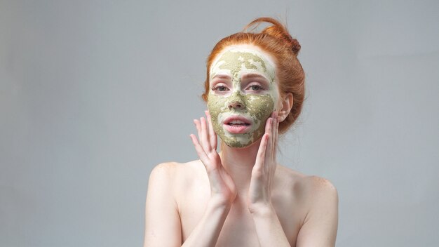 Een jonge vrouw met een groen droog moddermasker op haar gezicht op een geïsoleerde muur