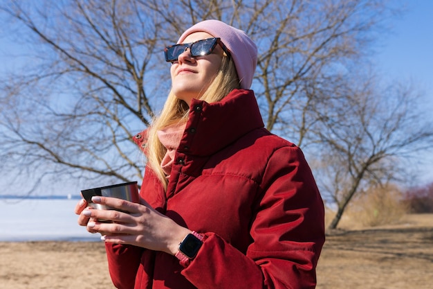 Een jonge vrouw met een hoed en een rood jasje drinkt thee uit een thermoskan