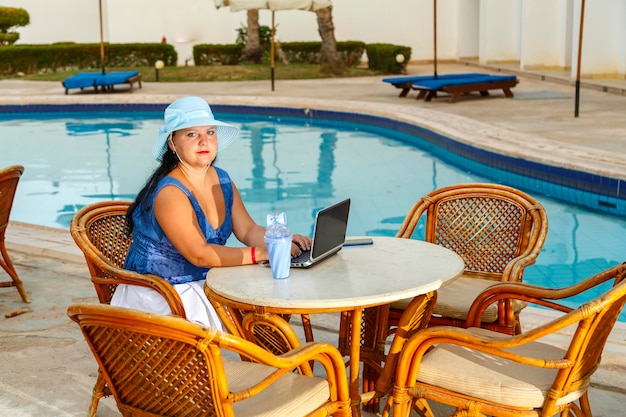 Een jonge vrouw met een hoed op aan een tafel in de ochtend naast het zwembad van het hotel