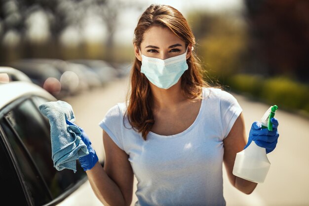 Een jonge vrouw met een masker op haar gezicht en beschermende handschoenen aan haar handen staat klaar om haar auto af te vegen met een doek in de ene en een fles met ontsmettingsmiddel in de andere hand.