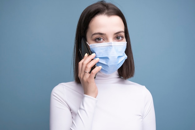 Een jonge vrouw met een medisch masker houdt een mobiele telefoon in haar handen en maakt een selfie op een blauwe achtergrond in de studio.