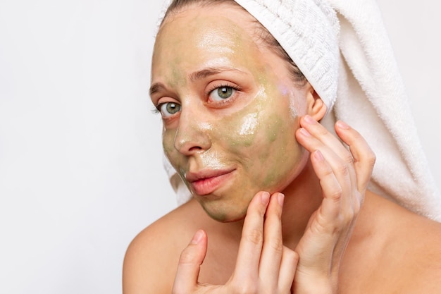 Een jonge vrouw met een witte handdoek op haar hoofd na een douche brengt een groen peelingmasker aan op het gezicht