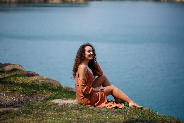 Een jonge vrouw met krullend haar en een glimlach op haar gezicht zit in een groene weide op een achtergrond van blauw meer. Warme zomerdag, gelukkig meisje, emoties van vreugde