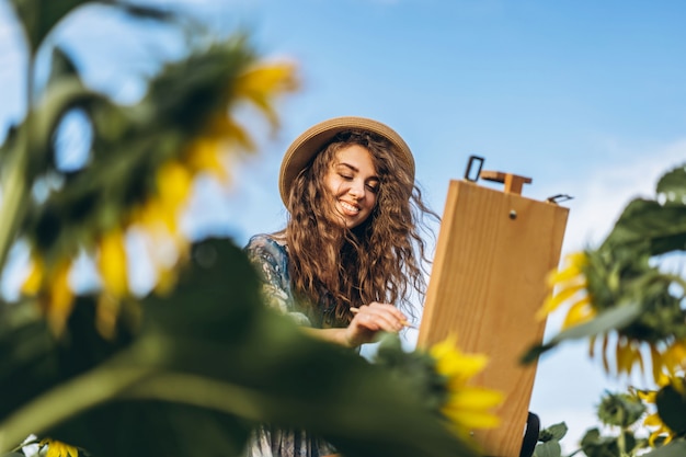 Een jonge vrouw met krullend haar en het dragen van een hoed schildert in de natuur. Een vrouw staat in een zonnebloem veld op een mooie dag