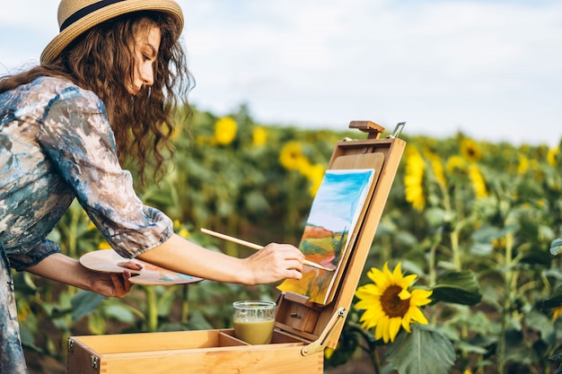 Een jonge vrouw met krullend haar en het dragen van een hoed schildert in de natuur. Een vrouw staat in een zonnebloem veld op een mooie dag