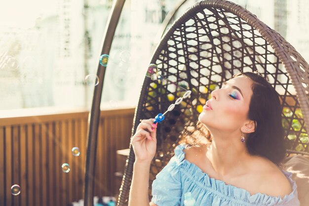 Een jonge vrouw met lichte make-up zit op een zomerterras in een hangstoel en blaast zeepbellen