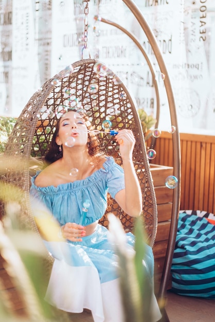 Een jonge vrouw met lichte make-up zit op een zomerterras in een hangstoel en blaast zeepbellen