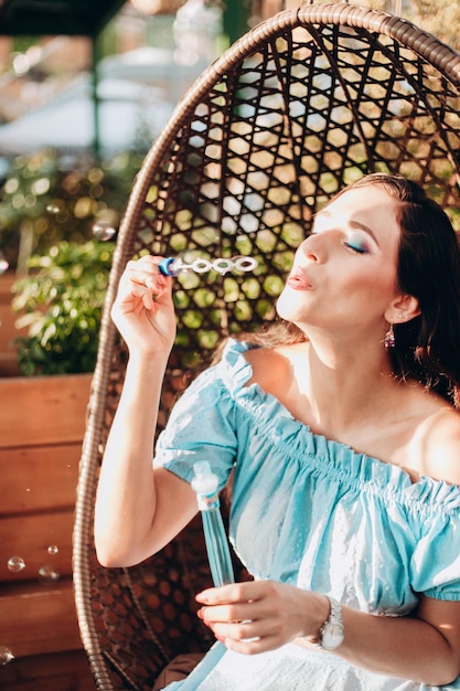 Een jonge vrouw met lichte make-up zit op een zomerterras in een hangstoel en blaast zeepbellen