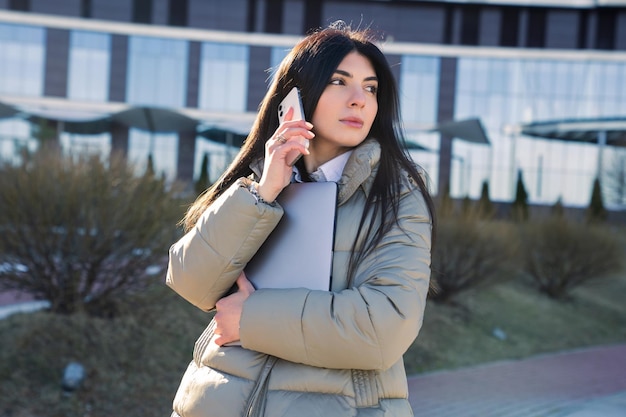 Een jonge vrouw praat aan de telefoon en houdt een laptop buiten vast